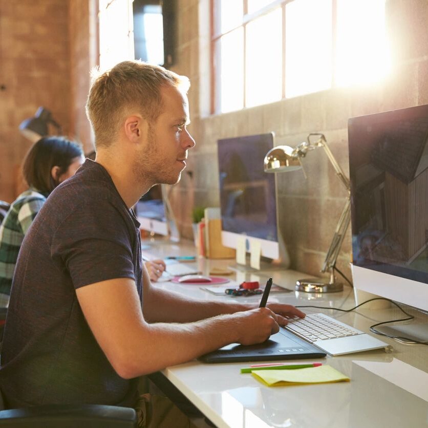 A man sits at a desk with two monitors in front of him.