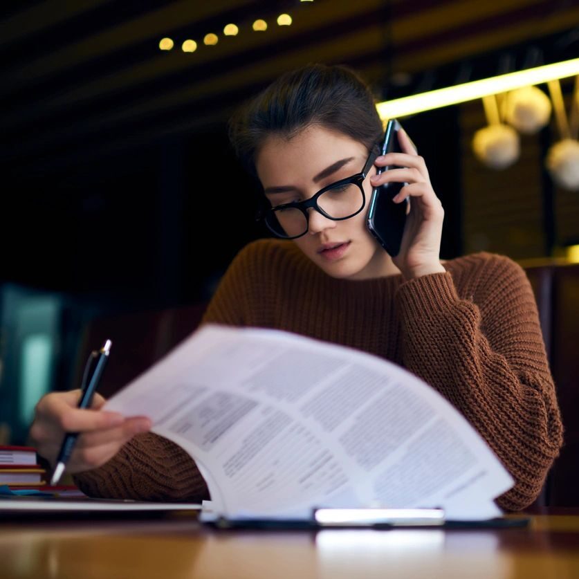 A woman is on the phone while sitting at a table with papers and a laptop, as she manages business development services.